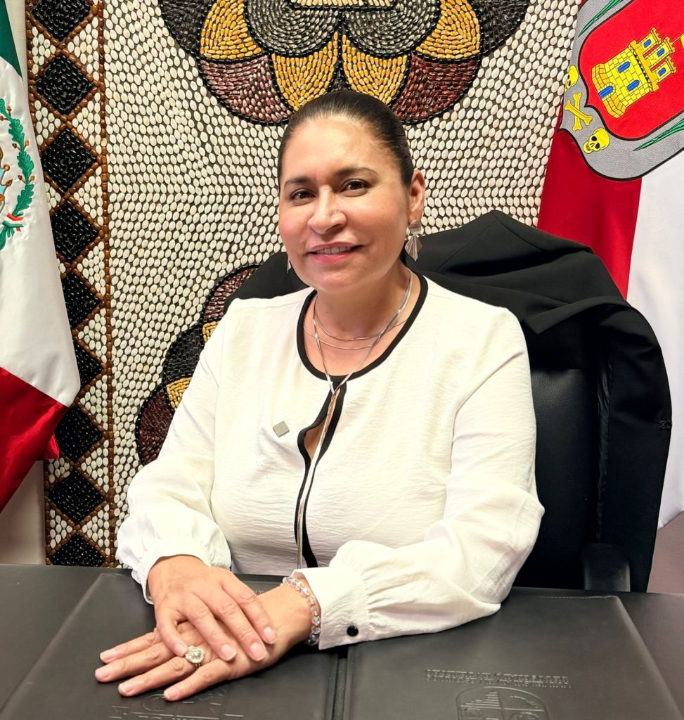 Una mujer sentada en una oficina con banderas de México y Tlaxcala al fondo, luciendo una blusa blanca y una expresión amigable.