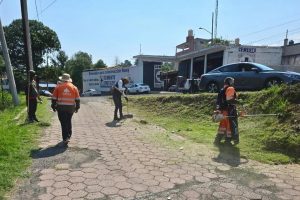 Caminando por una acera en una zona urbana, varias personas con equipo de protección realizan trabajos de mantenimiento o limpieza en un área pública, con edificios y vehículos en el fondo.