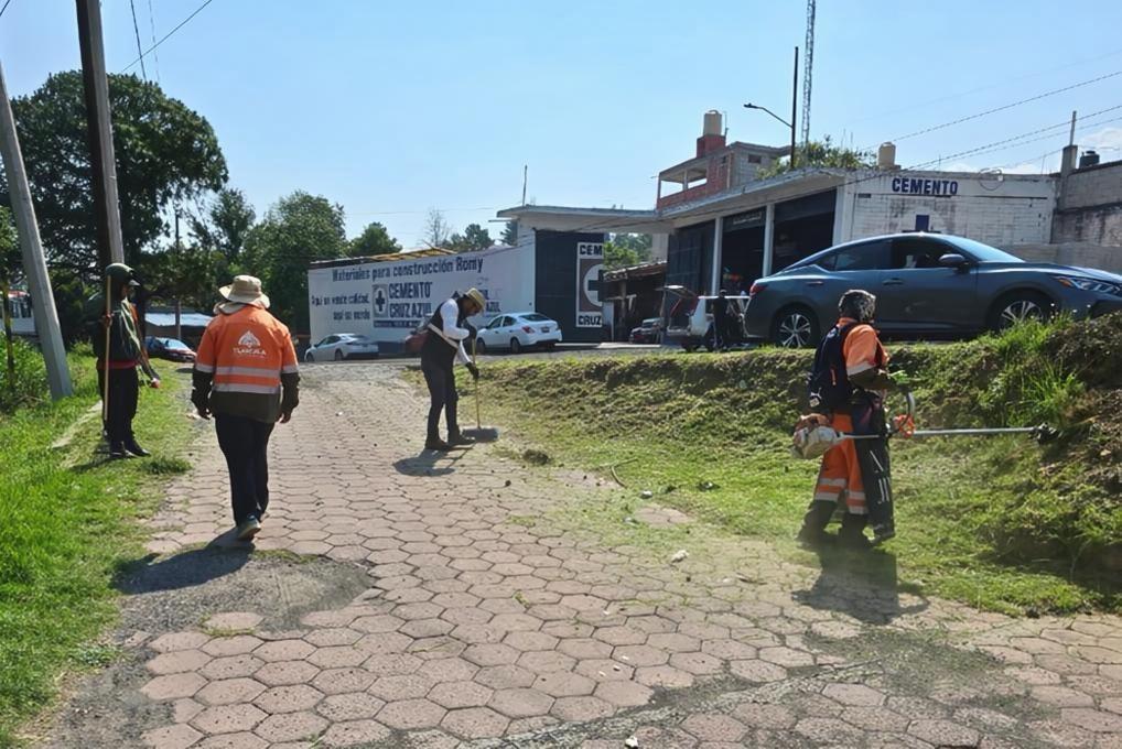 Caminando por una acera en una zona urbana, varias personas con equipo de protección realizan trabajos de mantenimiento o limpieza en un área pública, con edificios y vehículos en el fondo.