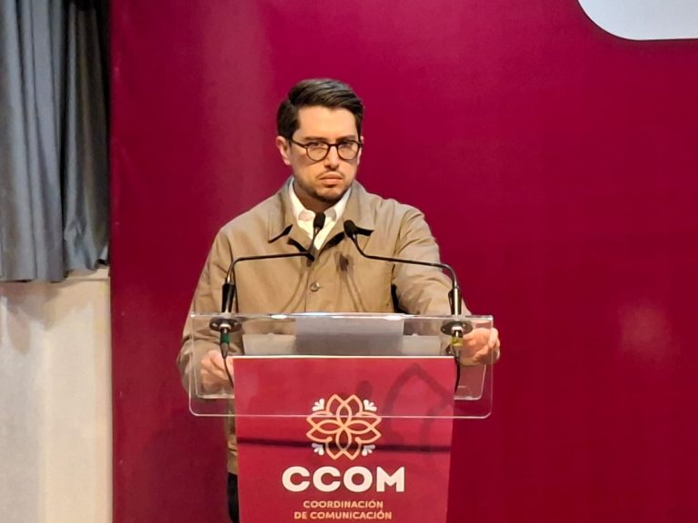 El hombre con gafas y chaqueta beige hablando en un podio con logotipo de la Coordinación de Comunicación en un evento formal.