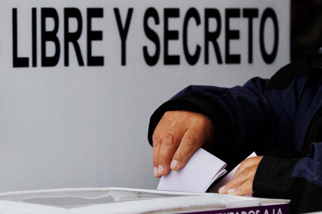 Una persona depositando un sobre en una urna electoral, con un cartel que dice "LIBRE Y SECRETO" en el fondo, en un proceso de votación.