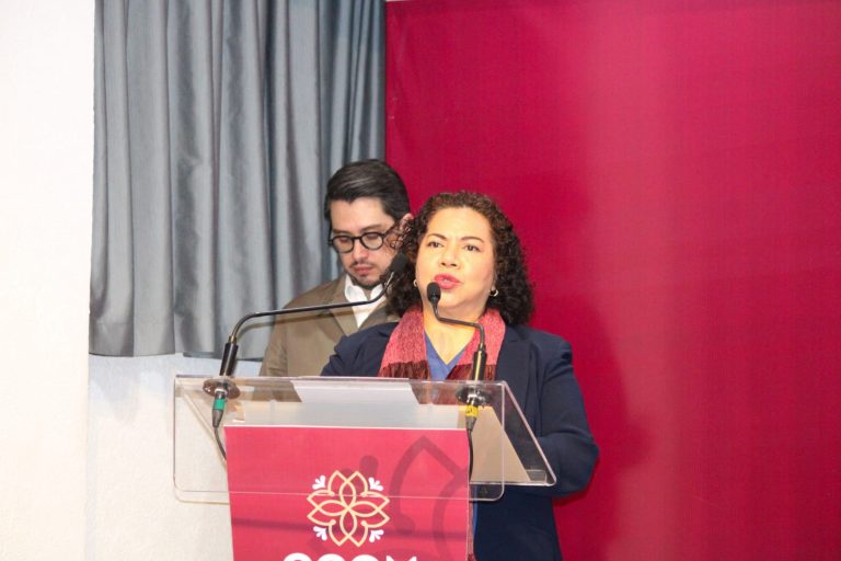 Mujer y hombre en un evento formal, la mujer habla en un micrófono en un podio con fondo rosa y cortinas grises.