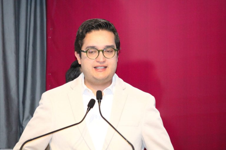 Joven hombre con gafas y cabello oscuro, vestido con saco blanco, hablando en un micrófono en un evento formal con fondo de cortinas grises y pared rosa.
