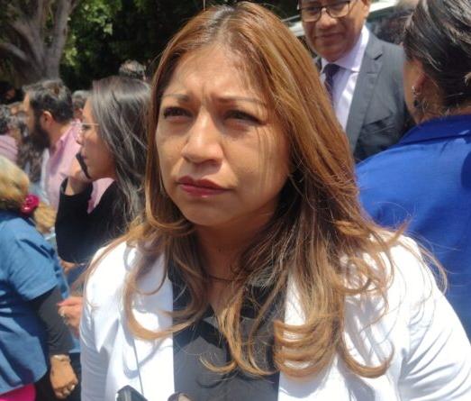 Una mujer con cabello castaño claro y expresión seria en un evento público, rodeada de varias personas, algunas con mascarillas y vestimenta formal, en un entorno al aire libre con árboles y luz natur.
