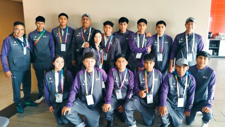 Atleta de Taekwondo en uniforme con medallas y medallones, acompañado por entrenadores y oficiales, en un evento deportivo en un espacio interior.