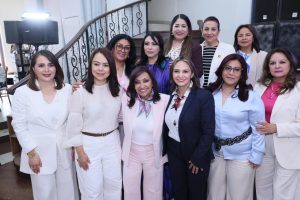 Grupo de mujeres profesionales en un evento formal, posando juntas en un espacio interior con escaleras y decoración moderna.