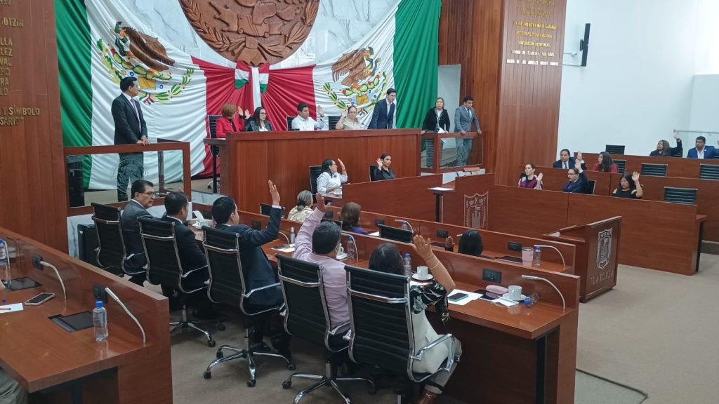 Un grupo de personas participa en una sesión en un recinto oficial con banderas de México y Tlaxcala en el fondo, en un ambiente formal y de discusión.