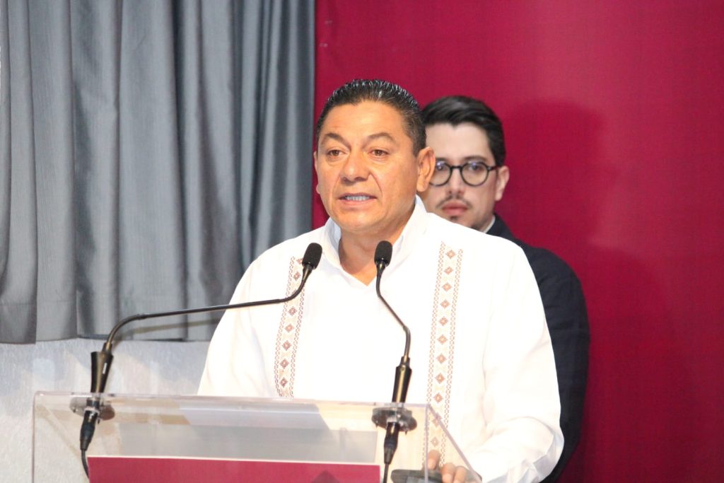 Hombre hablando en un evento formal con micrófonos, vestido con camisa blanca y fondo de cortinas grises y rojo.