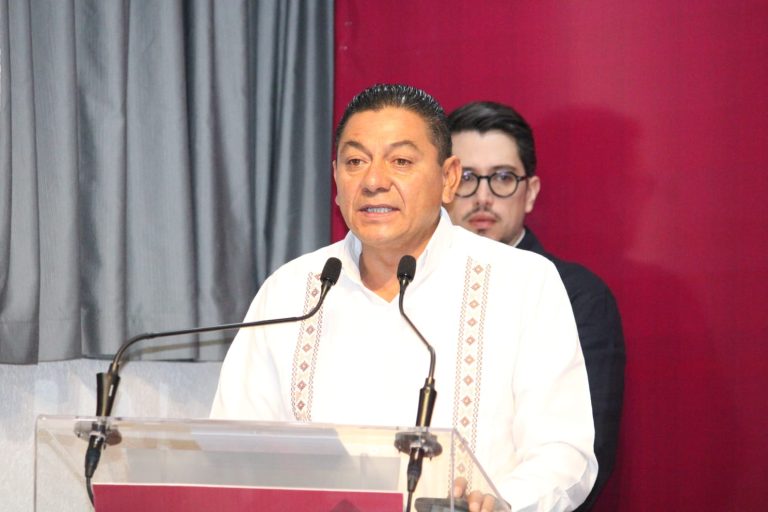 Hombre hablando en un evento formal con micrófonos, vestido con camisa blanca y fondo de cortinas grises y rojo.