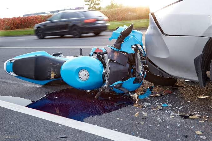 Motocicleta accidentada en la carretera tras chocar con un automóvil, con daños visibles y caída en el pavimento.