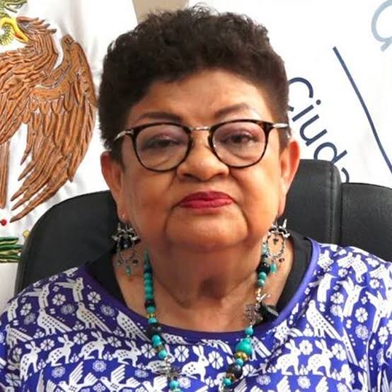 Mujer mayor con gafas y peinado corto, vestida con blusa azul con detalles blancos, en un entorno oficial con escudo y bandera mexicana al fondo.