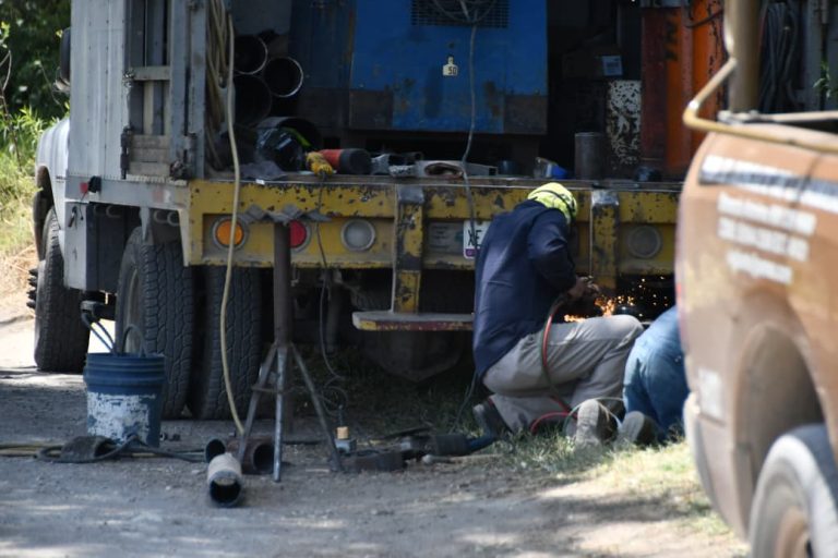 Cortador de metal trabajando en una obra, usando equipo de protección y herramientas, en un entorno exterior con un camión y otros vehículos presentes.