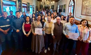 Grupo de personas en una ceremonia de reconocimiento en un salón elegante con pinturas y candelabros, celebrando logros y entregando certificados.