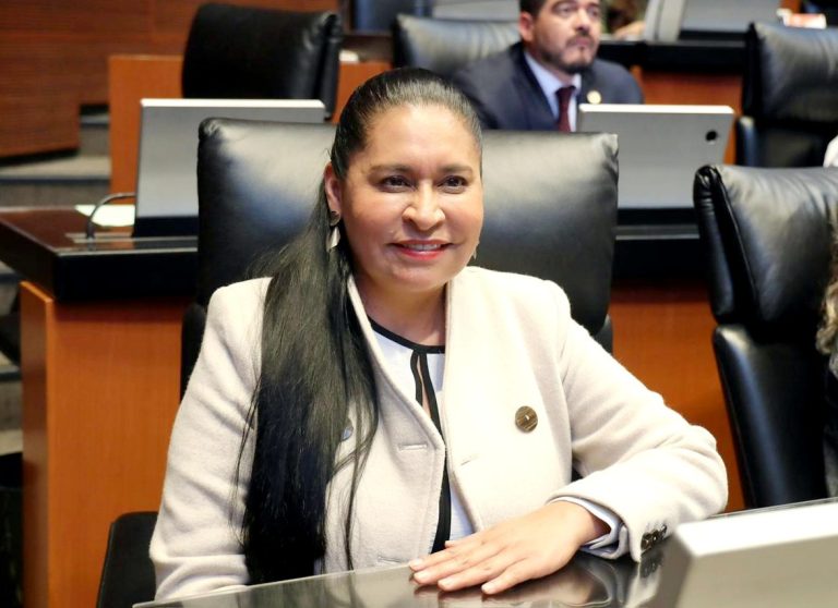 Mujer en una sala de reuniones, sonriendo y con cabello largo, vestida con blusa blanca y chaqueta beige, en un entorno formal con otros asistentes y computadoras.