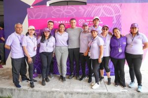 Grupo de personas en evento oficial en Tlaxcala, con fondo rosa y logotipo del gobierno estatal, participando en actividad comunitaria o institucional.