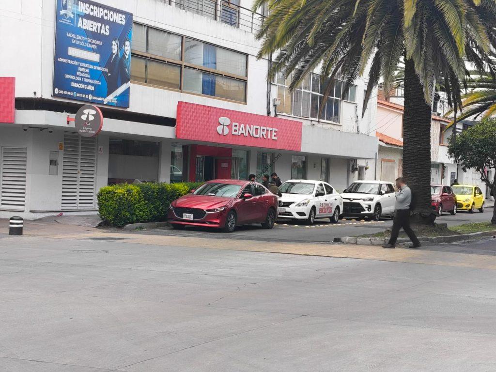 Tarjeta de crédito y sucursal bancaria Banorte frente a un edificio con palmeras y autos estacionados en la calle.