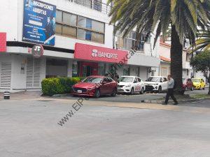 Tarjeta de crédito y sucursal bancaria Banorte frente a un edificio con palmeras y autos estacionados en la calle.
