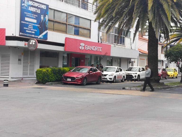 Tarjeta de crédito y sucursal bancaria Banorte frente a un edificio con palmeras y autos estacionados en la calle.