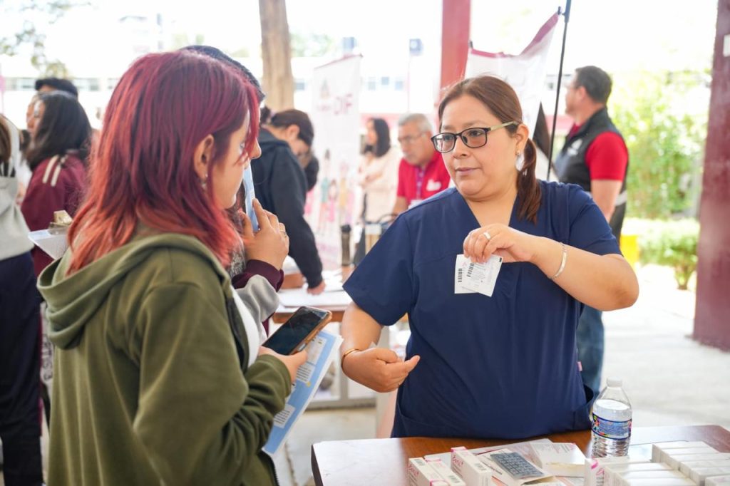 Mujer en un evento de salud entregando información y atendiendo a una asistente en un espacio abierto con varias personas en el fondo.