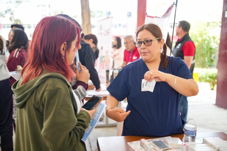 Mujer en un evento de salud entregando información y atendiendo a una asistente en un espacio abierto con varias personas en el fondo.