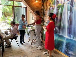 Una mujer entregando un certificado a una mujer en un espacio interior decorado con una cascada pintada en la pared y plantas naturales en la ventana.