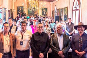 Grupo de personas en una sala con decoraciones clásicas, algunos con vestimenta formal y otros con ropa casual, en un evento o reunión social en un espacio elegante.
