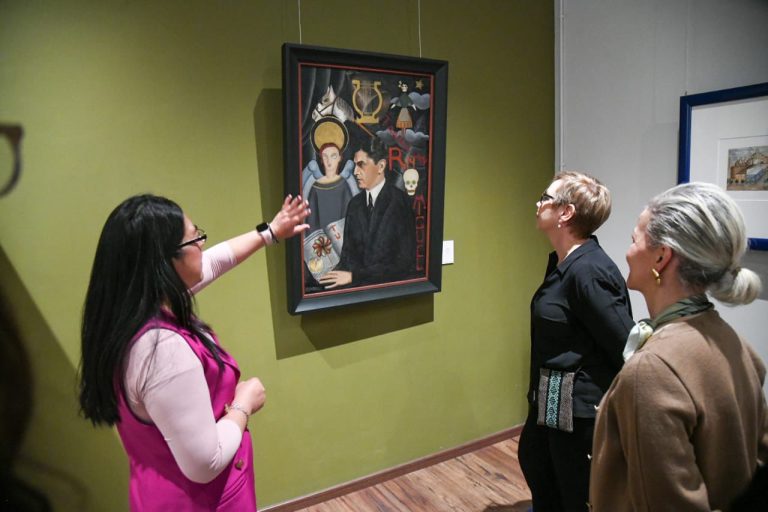 Una mujer explica una obra de arte a dos visitantes en una galería, mientras observan atentamente la pintura enmarcada en la pared de un espacio cultural.