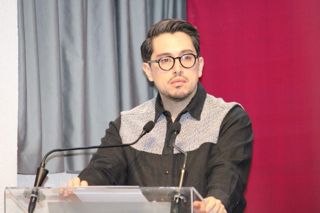 Hombre con gafas y barba, hablando en un podio durante una conferencia o evento formal.