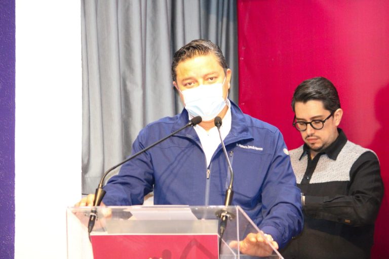 Hombre con mascarilla en un podio, hablando en un evento formal con fondo de cortinas grises y rojas.