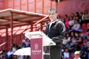 Persona hablando en un podio durante un evento público en un espacio abierto con audiencia en el fondo, en Tlaxcala, México.
