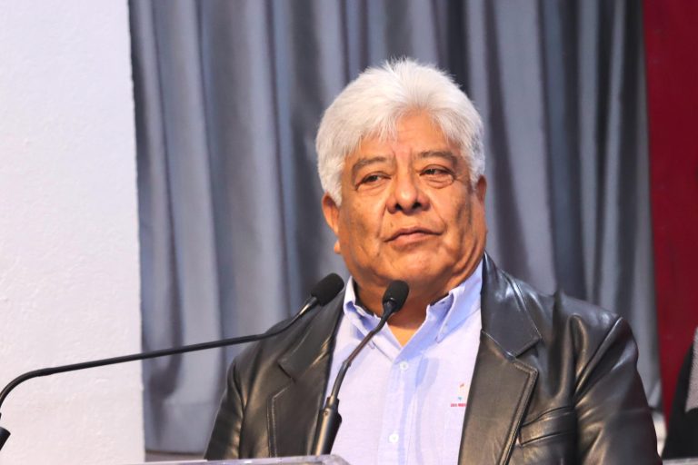 Un hombre mayor con cabello gris y vestimenta formal, hablando en un micrófono durante un evento oficial, con fondo de cortinas grises y bandera mexicana parcialmente visible.