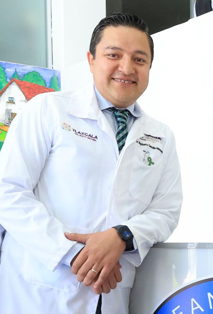 Médico profesional con bata blanca, sonriendo y posando en consulta, con fondo de pintura colorida de una casa y árboles.