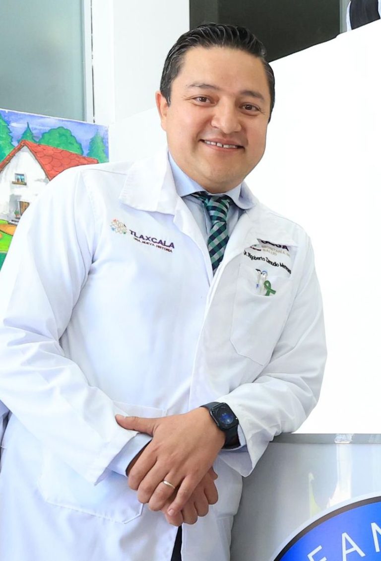 Médico profesional con bata blanca, sonriendo y posando en consulta, con fondo de pintura colorida de una casa y árboles.