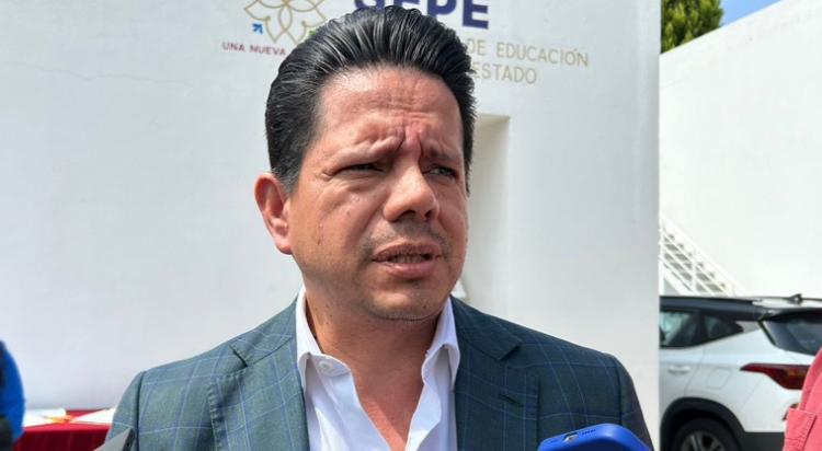 Hombre hablando en una entrevista al aire libre, vestido con traje y camisa blanca, en un entorno urbano con un edificio y un coche visible en el fondo.
