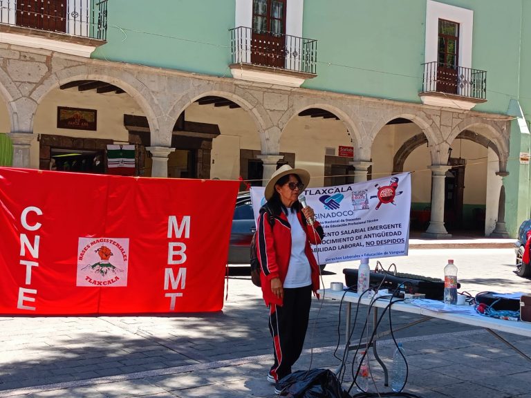 Una mujer con sombrero y ropa deportiva participa en una manifestación en Tlaxcala, con banderas y pancartas que exigen justicia social y derechos laborales en un espacio público con arquitectura colo.