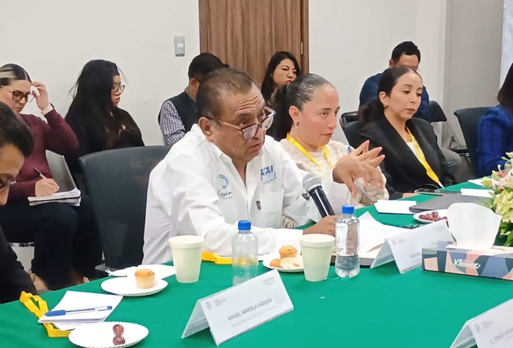 Persona en una reunión formal discutiendo temas importantes, rodeada de asistentes atentos en un entorno de oficina.