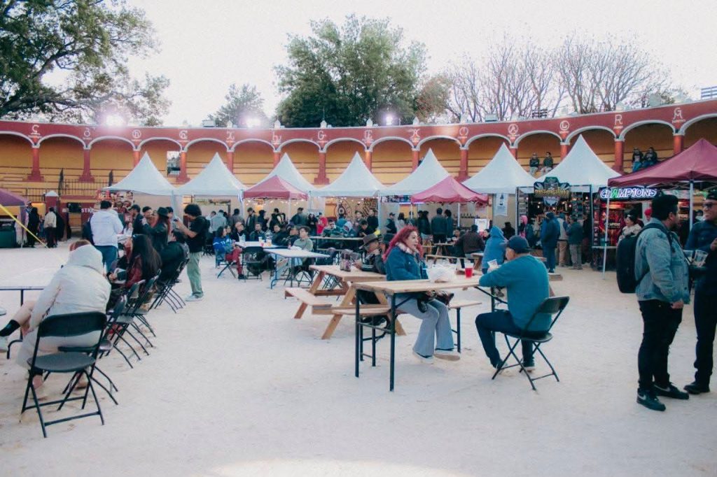 Evento al aire libre con mesas, sillas y puestos de comida en un espacio abierto con estructura arquitectónica colorida y árboles en el fondo.