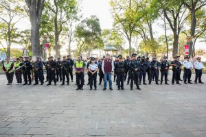 Grupo de policías y oficiales en una plaza pública con árboles y farolas, en una reunión o evento oficial en Tlaxcala, México.