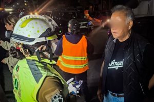Un oficial de policía revisa documentos a un hombre en una calle durante la noche, con otros oficiales y vehículos de emergencia en el fondo, en una escena que parece ser una intervención o control po.