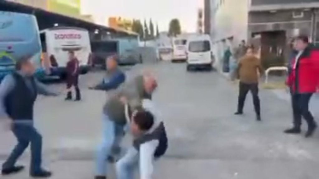 Personas peleando en la calle, algunos con movimientos agresivos, en un entorno urbano con autobuses y edificios al fondo.