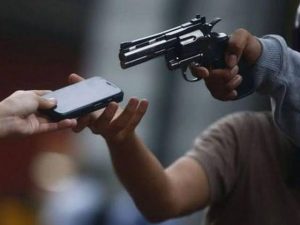 Un arma de fuego apuntando hacia un teléfono móvil en un entorno interior, con una mano sosteniendo el arma y otra mano sosteniendo el teléfono, en una situación que parece de tensión o amenaza.