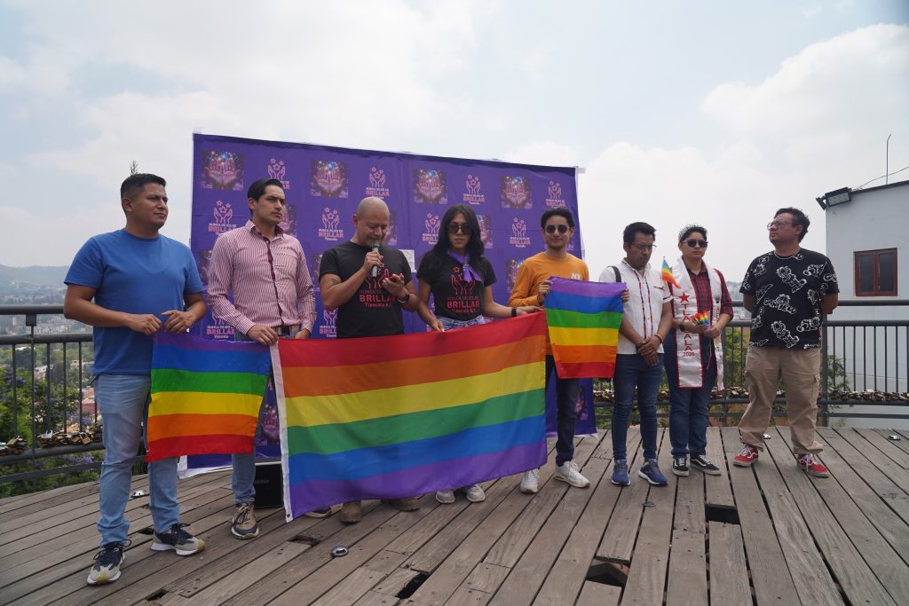 Bandera del orgullo LGBTQ+ en un evento al aire libre con varias personas en una plataforma de madera, algunas sosteniendo banderas arcoíris, en un día soleado con cielo parcialmente nublado.