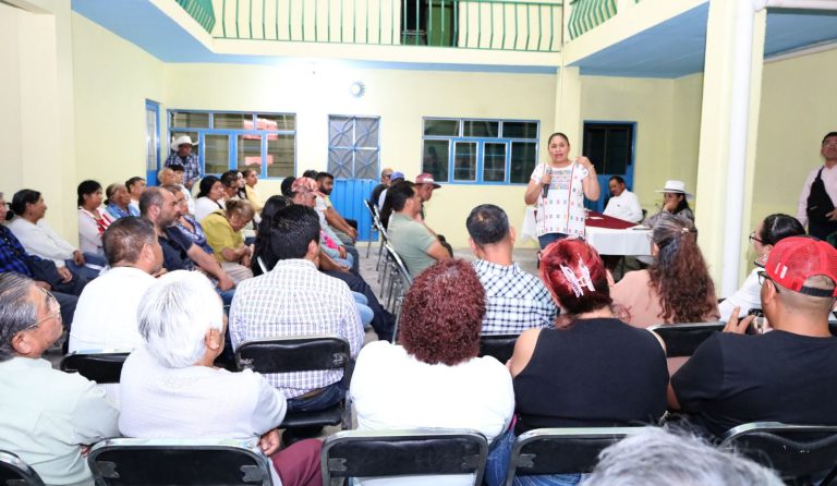 Asunto de una reunión comunitaria con una mujer hablando frente a un grupo de personas en un espacio cerrado.