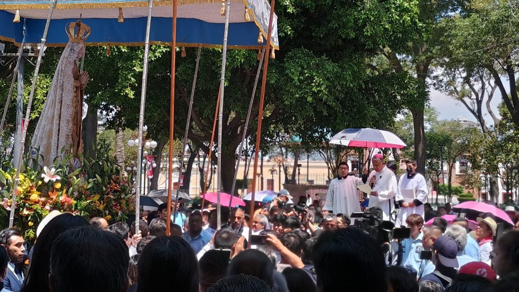Una procesión religiosa en Tlaxcala con una multitud de personas participando y sosteniendo paraguas en un día soleado. La imagen muestra una celebración tradicional con una figura religiosa en un alt.