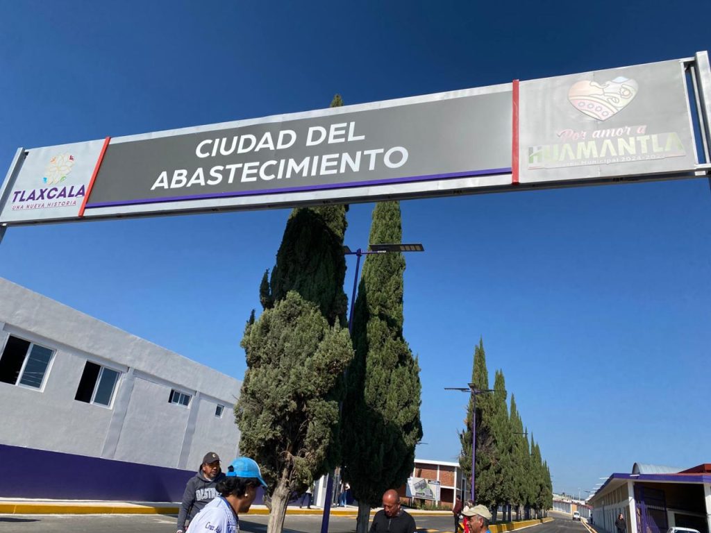 Barrera de entrada a la Ciudad del Abastecimiento en Tlaxcala, con árboles y edificios en el fondo, bajo un cielo despejado y azul.