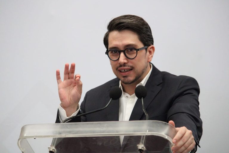 Un hombre con gafas y traje oscuro hablando en un podio con micrófonos, haciendo una señal con la mano durante una conferencia o discurso formal.