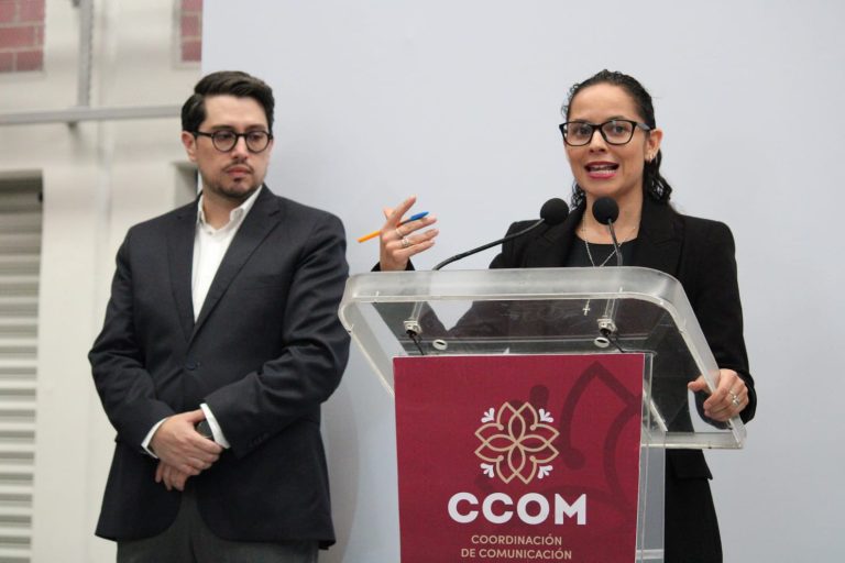 Dos personas en una conferencia, una mujer hablando en un podio con logotipo de CCOM, y un hombre observando atentamente, en un entorno profesional y formal.