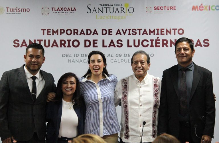 Grupo de cinco personas en una conferencia, sonriendo y posando frente a un cartel que anuncia la temporada de avistamiento en el Santuario de las Luciérnagas en Tlaxcala.