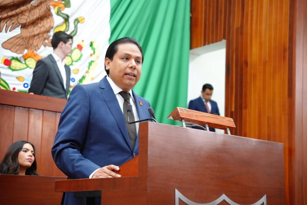 Hombre en traje azul hablando en un podio durante una ceremonia oficial en un recinto con bandera de México y fondo de madera.