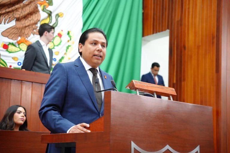Hombre en traje azul hablando en un podio durante una ceremonia oficial en un recinto con bandera de México y fondo de madera.